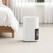 좌측에 원목 프레임 침대의 일부가 보이고, 베이지색 러그가 깔린 바닥 중앙에 흰색 LG 휘센 제습기 본체가 똑바로 세워져 있는 실내 이미지입니다. 제품 뒤쪽으로는 흰색 시폰 커튼이 쳐진 창문이 보입니다.