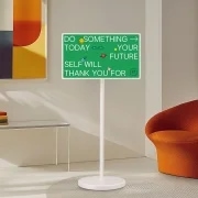 초록색 바탕에 흰색 글씨로 "DO SOMETHING → TODAY → YOUR FUTURE SELF WILL THANK YOU FOR"라고 적힌 간판이 흰색 기둥에 붙어 있고, 옆에는 주황색 의자와 테이블, 액자가 있는 사진.