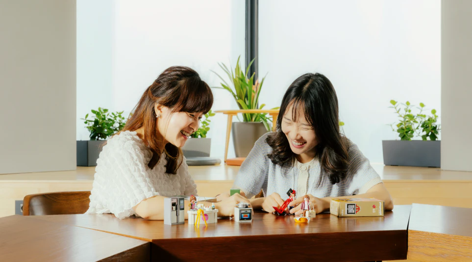 LG전자 플레이모빌을 기획한 김혜진 책임과 이지원 선임이 책상 위에 비치되어 있는 플레이모빌 피규어 세트들을 조작하며 환하게 웃고 있다.
