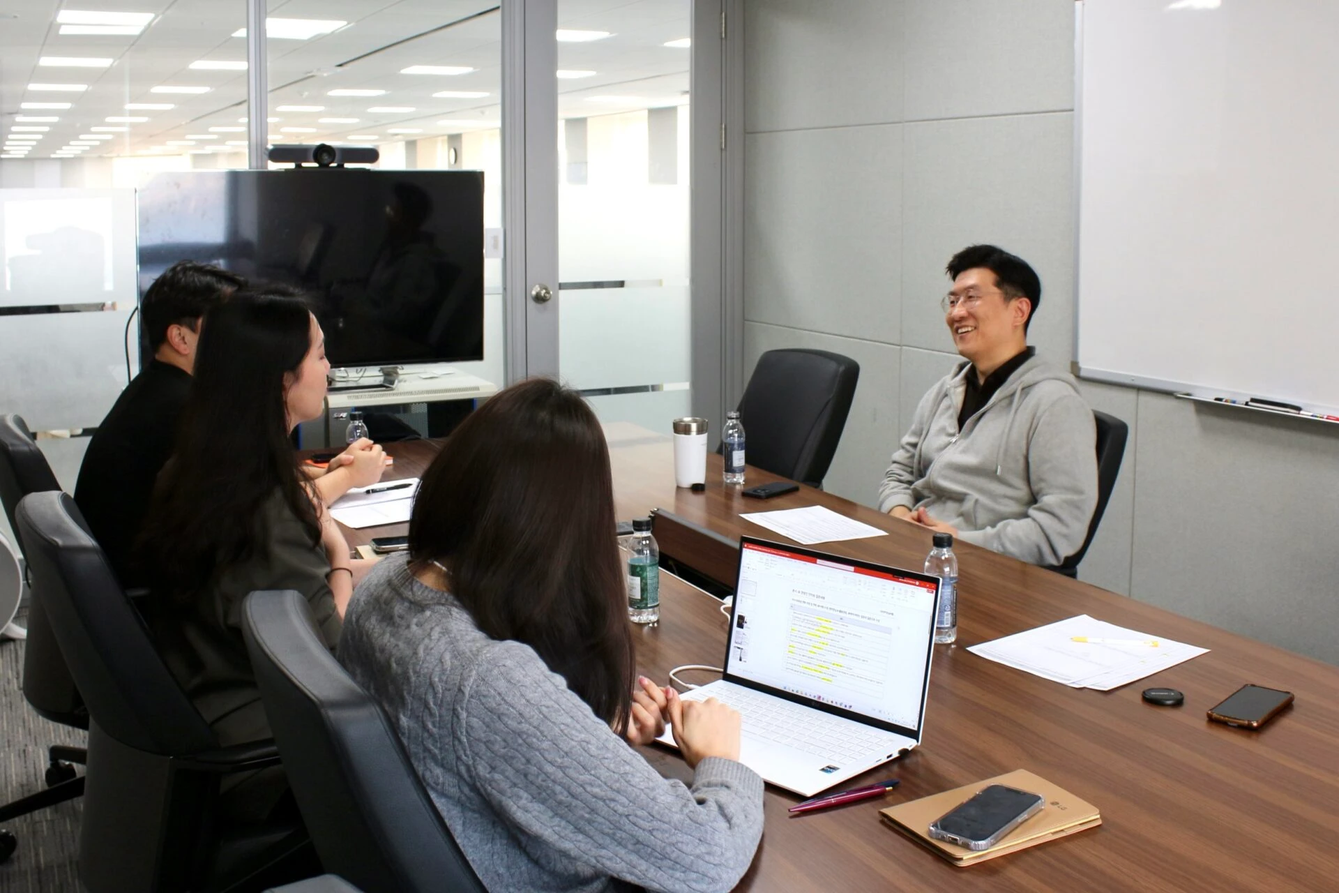 DX센터의 센터장 조정범 전무와 담당자들이 회의실에서 대화를 나누는 모습 회의실 안에서 네 명의 담당자들이 테이블을 사이에 두고 마주보고 앉아 있다. 조정범 센터장이 회의실 벽 쪽 좌석에 앉아 웃고 있으며, 나머지 세 명은 반대편에 앉아 메모하거나 노트북을 사용하고 있다. 테이블 위에는 종이, 스마트폰, 텀블러, 생수병, 펜 등이 놓여 있다. 회의실 안쪽 벽에는 화이트보드가 부착되어 있고, 유리문 너머에는 오픈 오피스 공간과 켜진 형광등이 보이며, TV 모니터와 화상 회의용 카메라도 함께 배치되어 있다. 참가자 모두는 캐주얼한 복장을 하고 있다.