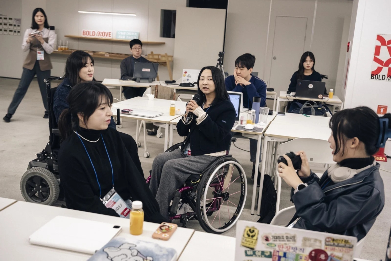 장애와 관계없이 다양한 사람들이 함께 모여 이야기하고 있는 포용적인 커뮤니티 모임 현장. 한 참여자가 마이크를 들고 진지하게 발언하고 있다.