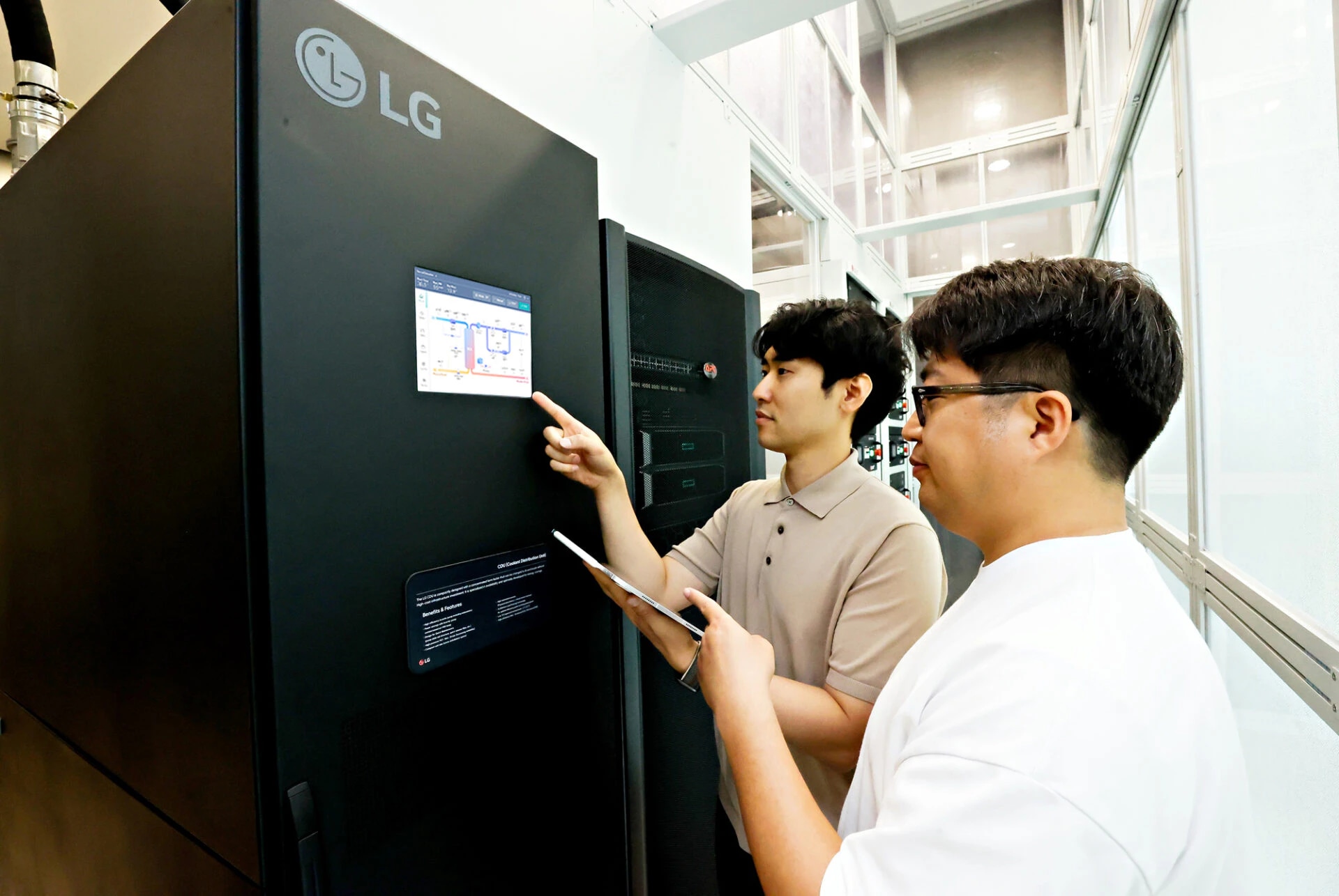 LG유플러스 직원이 AI 데이터센터 액체 냉각 솔루션인 LG전자 CDU를 테스트하고 있다.(사진 출처: LG유플러스) 기기 전면에는 LG 로고와 터치 디스플레이가 있으며, 한 남성이 화면을 손가락으로 가리키고 다른 한 남성은 태블릿을 들고 서 있다. 기기 측면에는 설명용 문구가 적힌 패널이 부착되어 있으며, 배경에는 유리벽과 파이프 설비, 다른 기기들이 보인다. 두 사람 모두 캐주얼한 복장을 하고 있다.