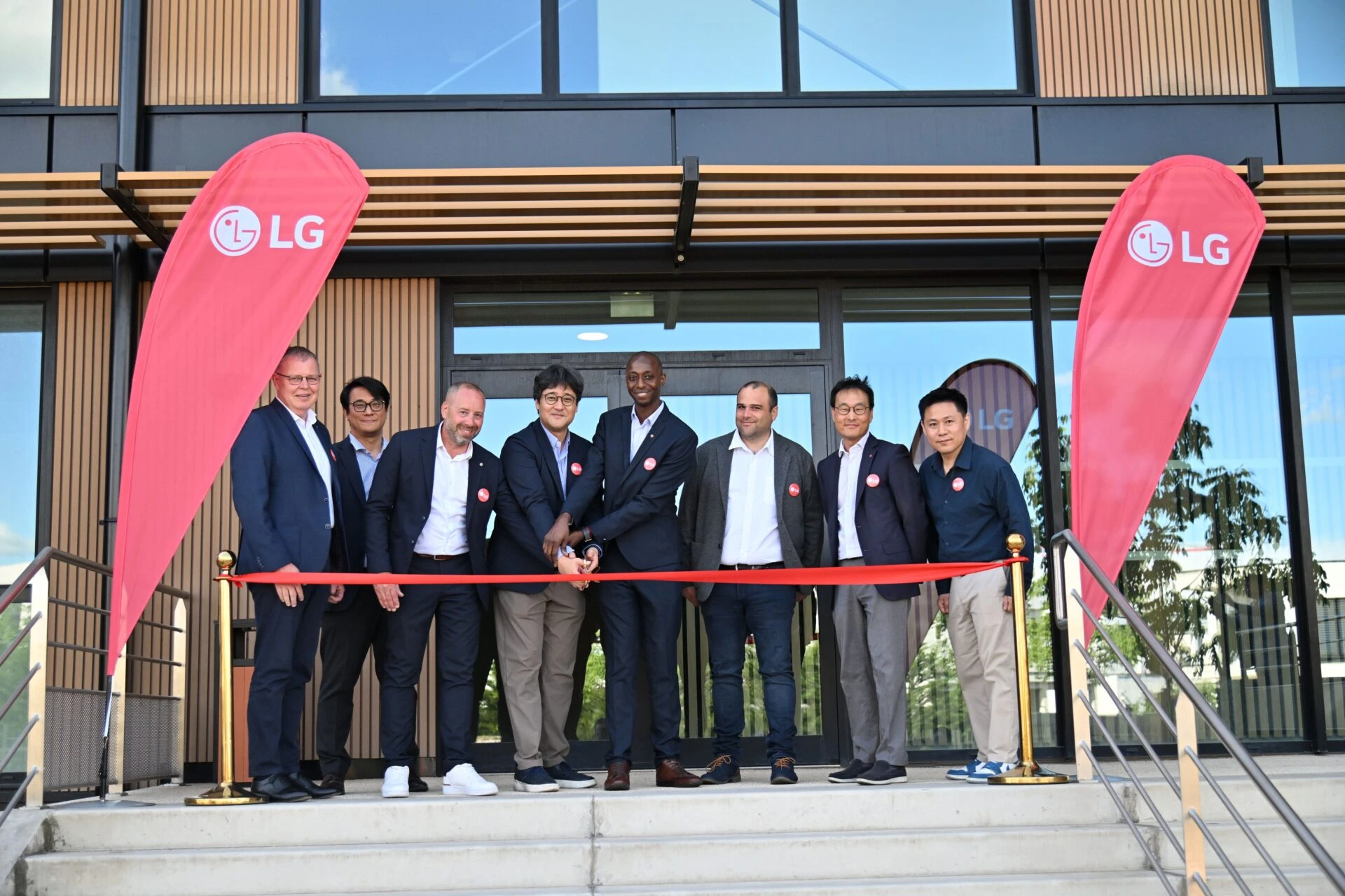 LG전자 프랑스 리옹 HVAC 아카데미 오픈식
LG 로고가 새겨진 붉은색 배너 깃발 두 개 사이에서 오픈식을 진행하고 있다.
총 아홉 명의 정장 차림 남성들이 건물 입구 앞에 나란히 서 있으며, 가운데 두 사람이 함께 빨간색 리본을 자르는 가위질을 하고 있다.
모두 가슴에 동일한 원형 배지를 달고 있으며, 밝게 웃거나 포즈를 취하고 있다.
건물 외관은 나무 패널과 유리창으로 구성되어 있고, 입구 유리문 안쪽에는 LG 로고가 반사되어 보인다.
공식적인 오프닝 행사로 보이며, 다양한 국적의 관계자들이 함께 자리한 모습이다.