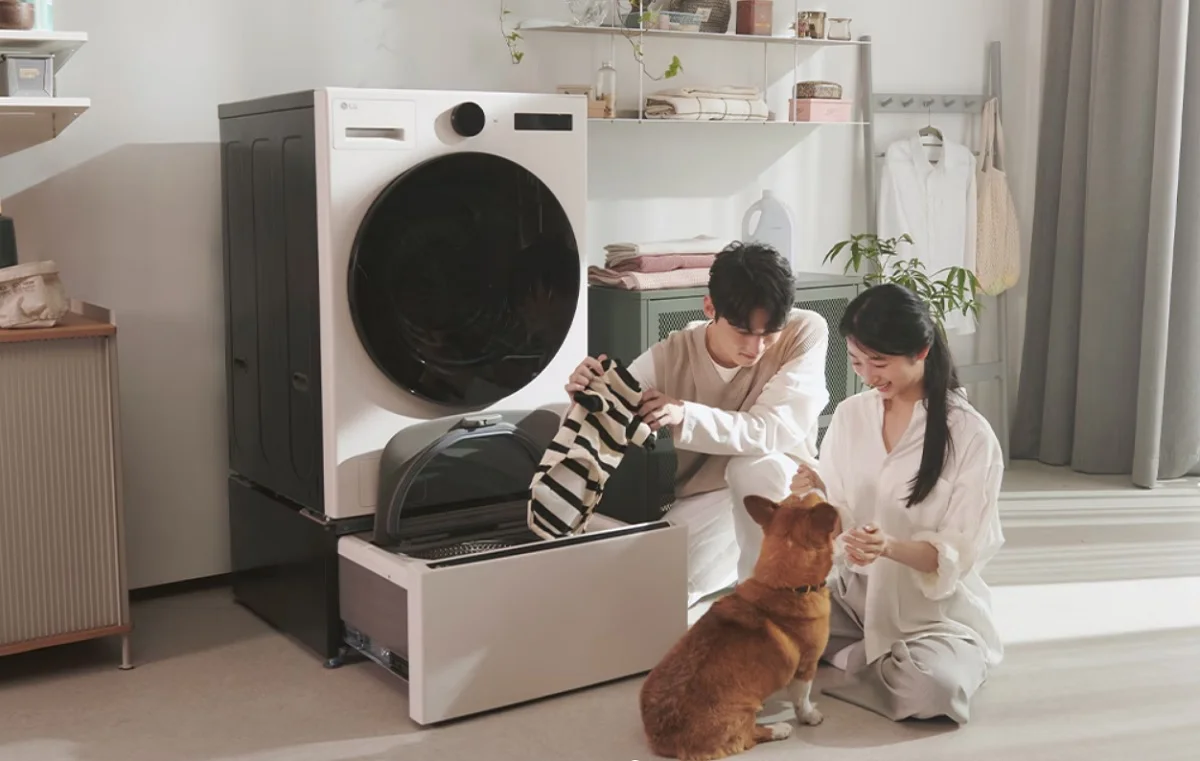 밝고 따뜻한 분위기의 실내에서 세탁기를 사용하는 모습. 세탁기 하단의 미니워시 서랍이 열려 있고, 남성이 스트라이프 무늬의 반려동물 옷을 넣으려는 모습을 보인다. 옆에 앉아 있는 여성은 반려견을 쓰다듬으며 웃고 있다. 세탁기 주변에는 선반과 식물, 생활용품들이 정돈돼 있어 아늑한 생활 공간을 연출한다.