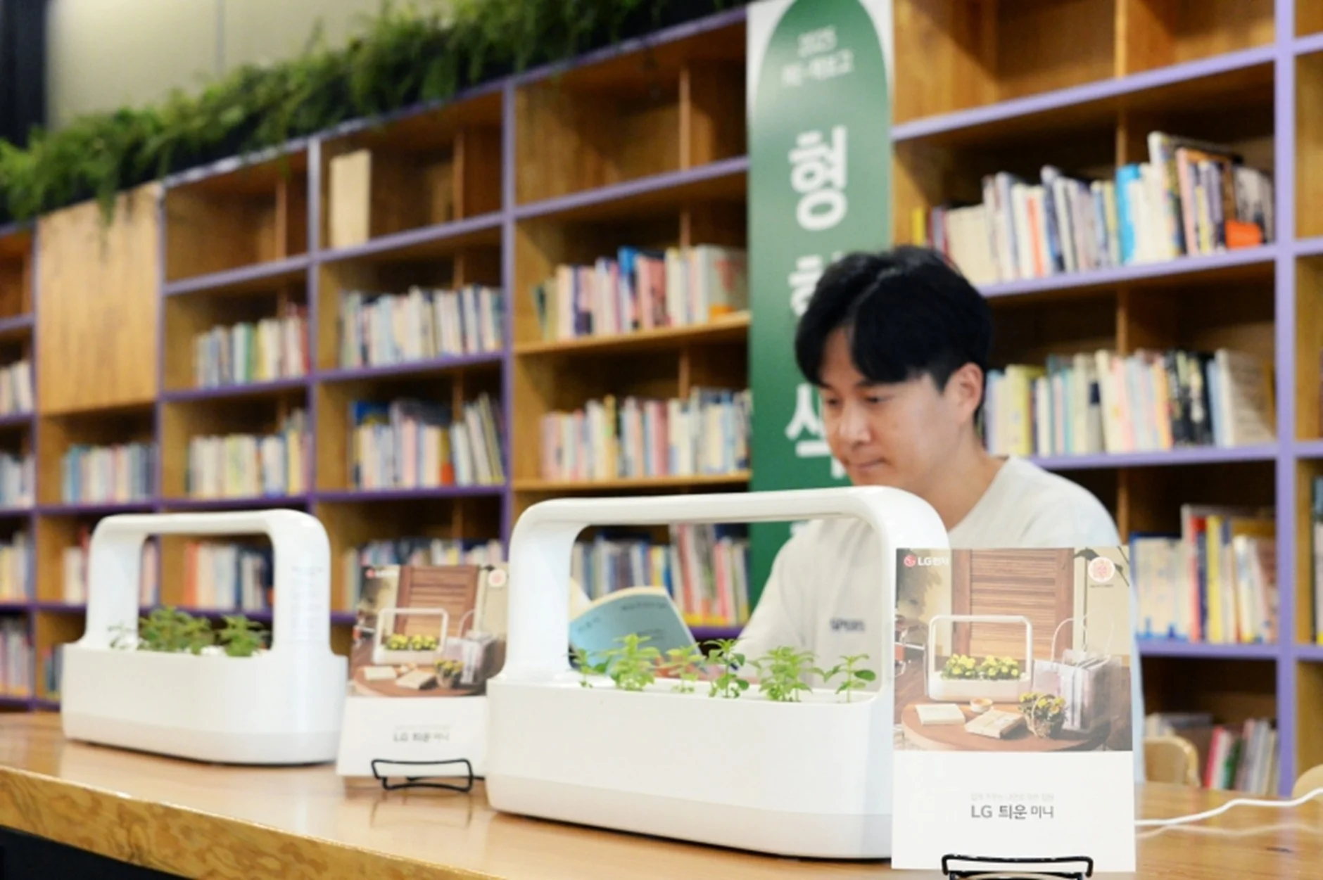 도서관으로 보이는 공간에서 한 남성이 책을 읽고 있다. 그의 앞 탁자에는 LG전자 식물 재배기 ‘LG 틔운 미니’ 제품 3대가 놓여 있으며, 각각에는 작게 자란 식물들이 심어져 있다. 제품 앞에는 홍보용 안내판이 세워져 있고, 배경에는 다양한 책이 꽂힌 책장이 가득하다. 위쪽에는 초록색 식물들이 천장 가까이 설치되어 있고, 가운데에는 ‘2025 희망서울 청년 책방’이라고 적힌 배너가 보인다. 전체적으로 차분하고 친환경적인 분위기를 자아낸다.