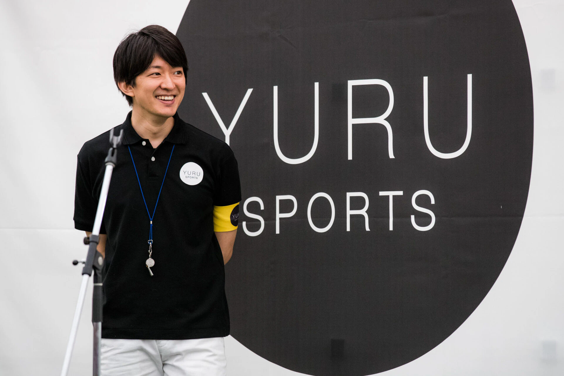 검은색 폴로 셔츠를 입고, 팔에 노란색 밴드를 착용한 남성이 흰색 YURU SPORTS 로고가 새겨진 큰 검은색 원형 배경 앞에 서 있다.  