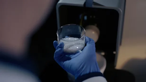 파란 장갑을 낀 연구원이 마린 글라스가 담긴 유리 잔을 손에 들고 있으며, 배경에는 실험 장비가 흐릿하게 보인다.