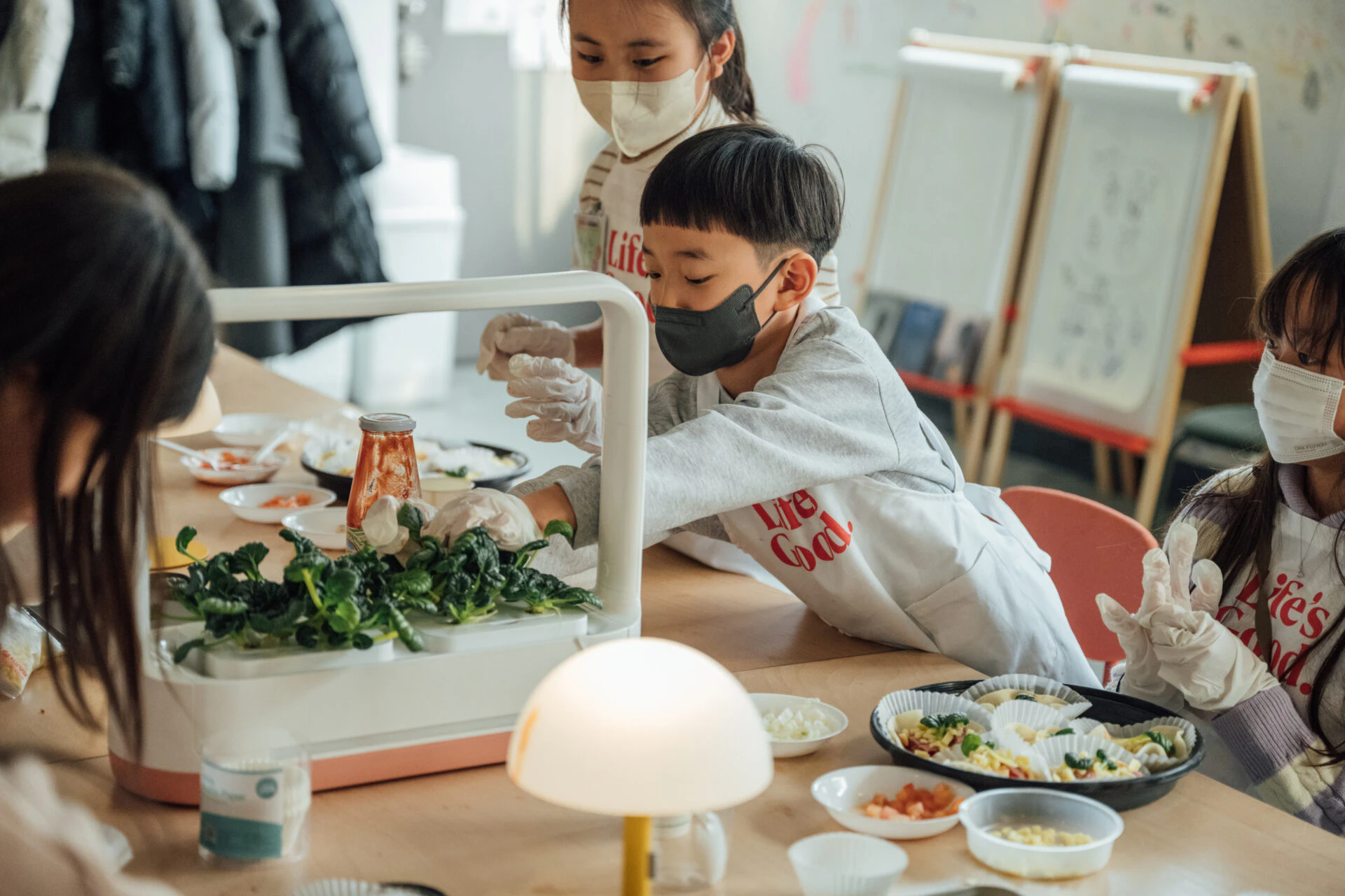 아이들이 마스크와 위생 장갑을 착용한 채 테이블에 앉아 요리 활동을 하고 있다. 한 아이는 작은 텃밭 모형에서 채소를 뽑고 있고, 다른 아이들은 주변에 놓인 재료를 만지고 있다.