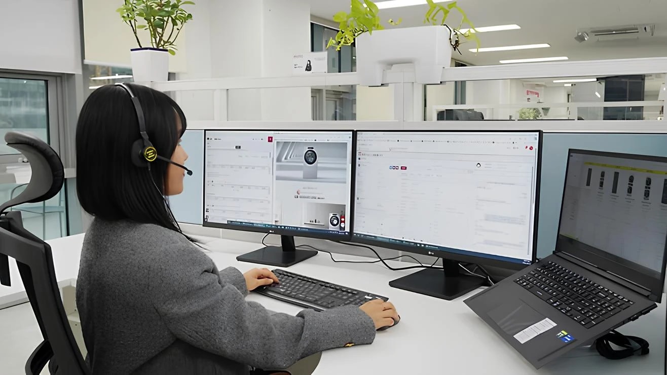 사무실에서 헤드셋을 착용한 여성이 듀얼 모니터와 노트북을 사용하며 업무를 보고 있는 모습. 모니터에는 제품 관련 웹페이지와 문서가 열려 있고, 책상 위에는 키보드와 마우스가 놓여 있으며, 칸막이 위에는 화분이 놓여 있다.