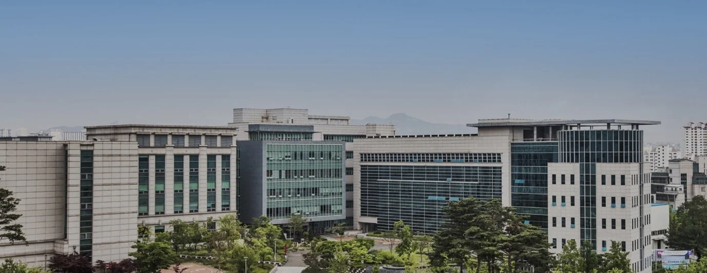 맑은 하늘 아래 촬영된 고려대학교 캠퍼스 전경 사진. 여러 동의 현대적인 회색·유리 외관 건물이 나란히 서 있고, 앞쪽에는 나무와 녹지가 보인다. 대학 건물들이 넓게 펼쳐진 전경을 담은 이미지다.
